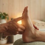 Mains d'un praticien en chemise claire massant délicatement le pied d'une personne dans une ambiance chaleureuse de spa.
